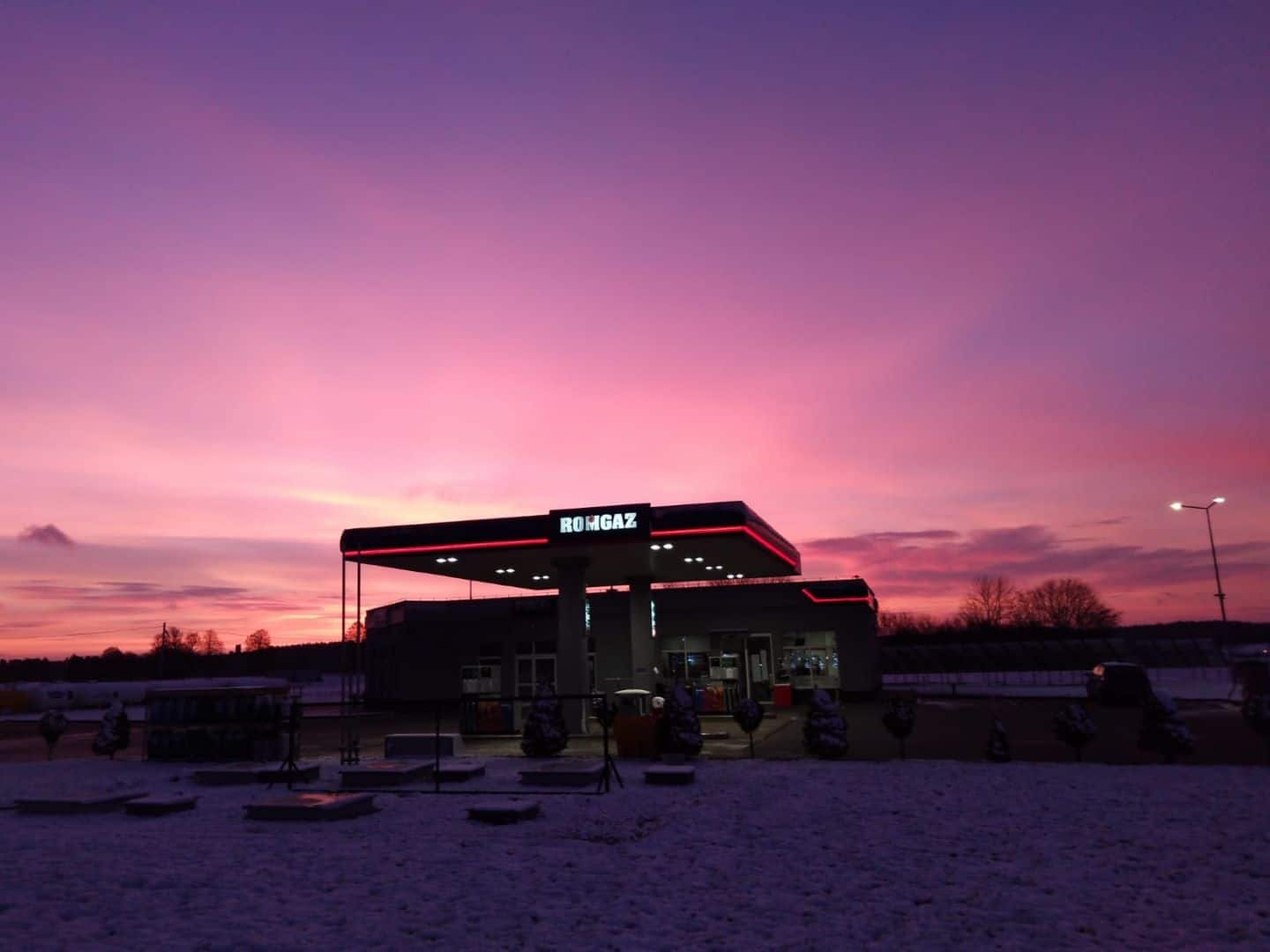 Stacja benzynowa pod tętniącym życiem różowym niebem zachodzącego słońca, ze śniegiem na ziemi i podświetlonym znakiem stacji.