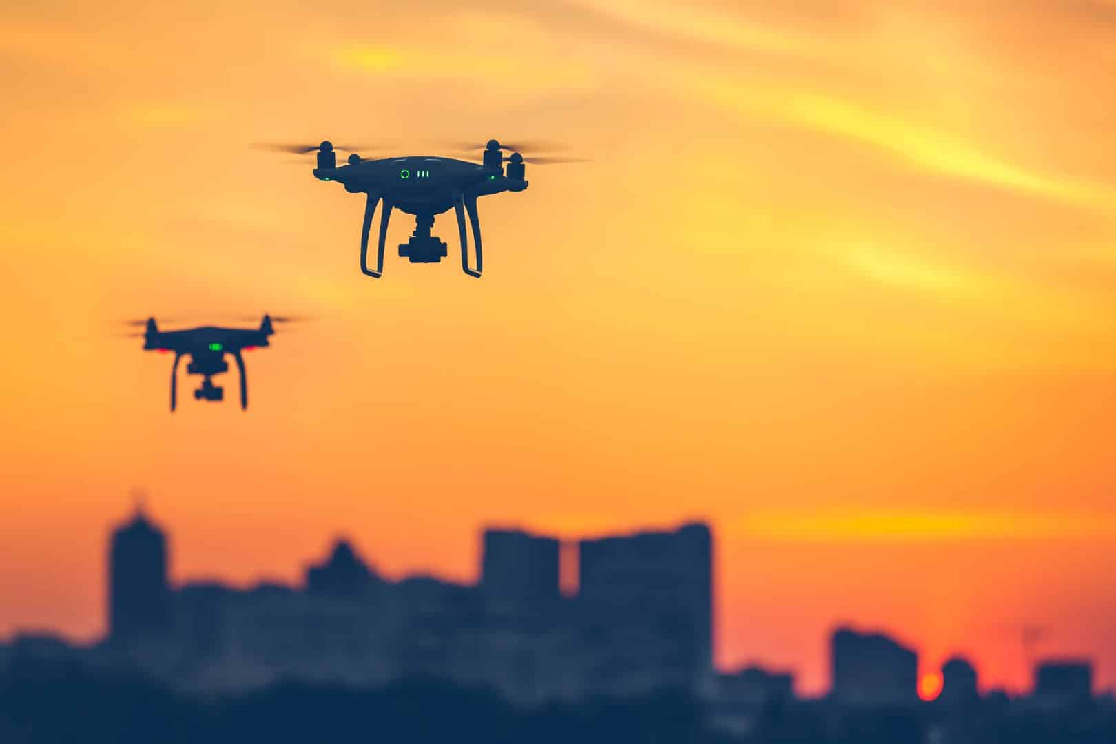 Dwa drony lecące na tle zachodzącego słońca z sylwetkami budynków miejskich w tle.