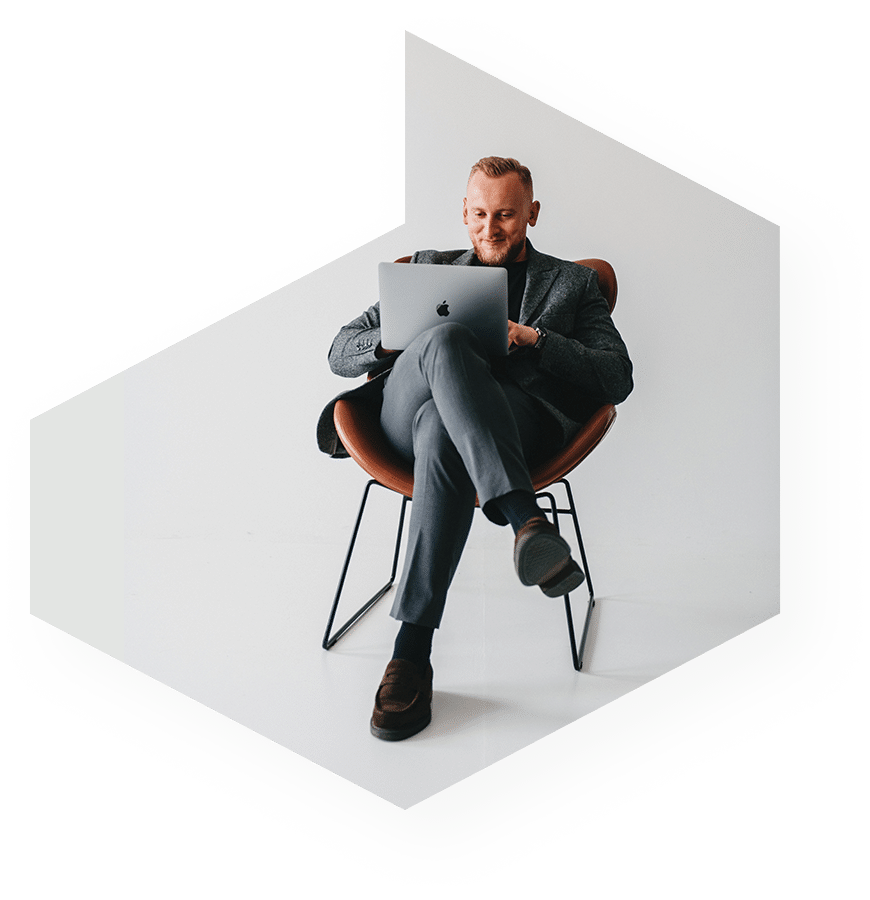 Mężczyzna w szarym garniturze siedzi wygodnie na nowoczesnym krześle i korzysta z laptopa. patrzy na ekran ze skupionym wyrazem twarzy, w minimalistycznej białej przestrzeni.