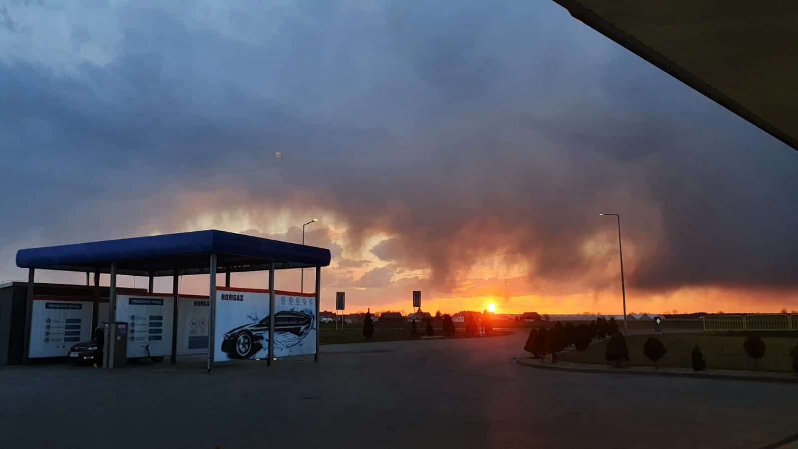 Stacja benzynowa pod ciemnym, pochmurnym niebem o zachodzie słońca. Słońce jest już blisko horyzontu, rzucając pomarańczowy blask. Stacja benzynowa ma niebieski baldachim z widocznymi pojazdami i oznakowaniem.