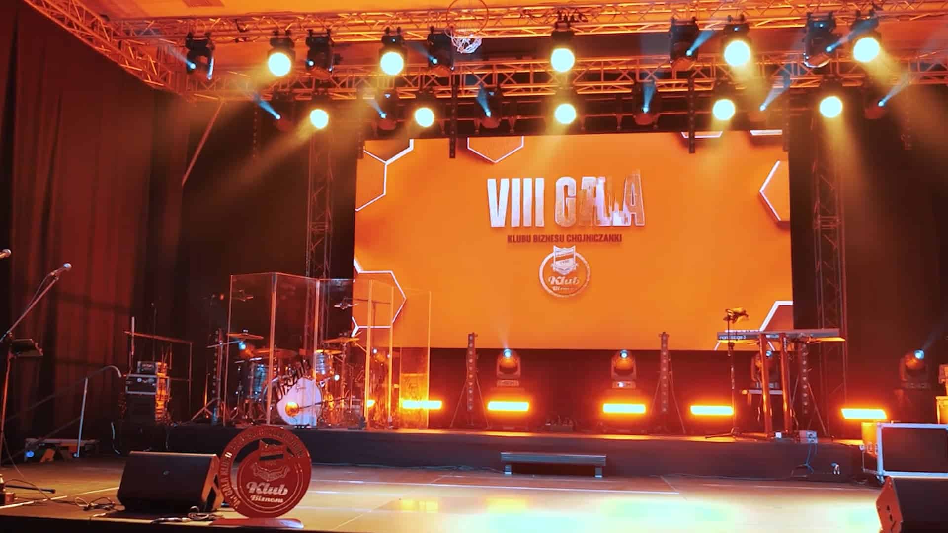 Oświetlona scena z zestawem perkusyjnym, mikrofonami, keyboardem i ekranem LED wyświetlającym "VIII GALA" pod pomarańczowym oświetleniem.