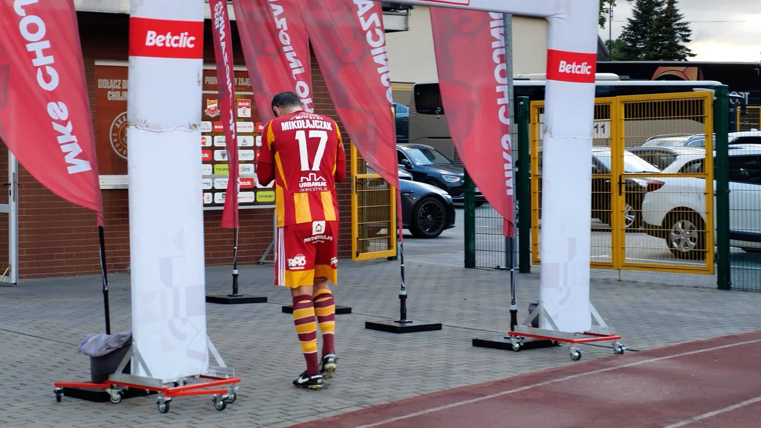 Piłkarz w koszulce z numerem 17 i czerwono-żółtymi paskami odchodzi pod banerami wydarzenia w pobliżu boiska sportowego.
