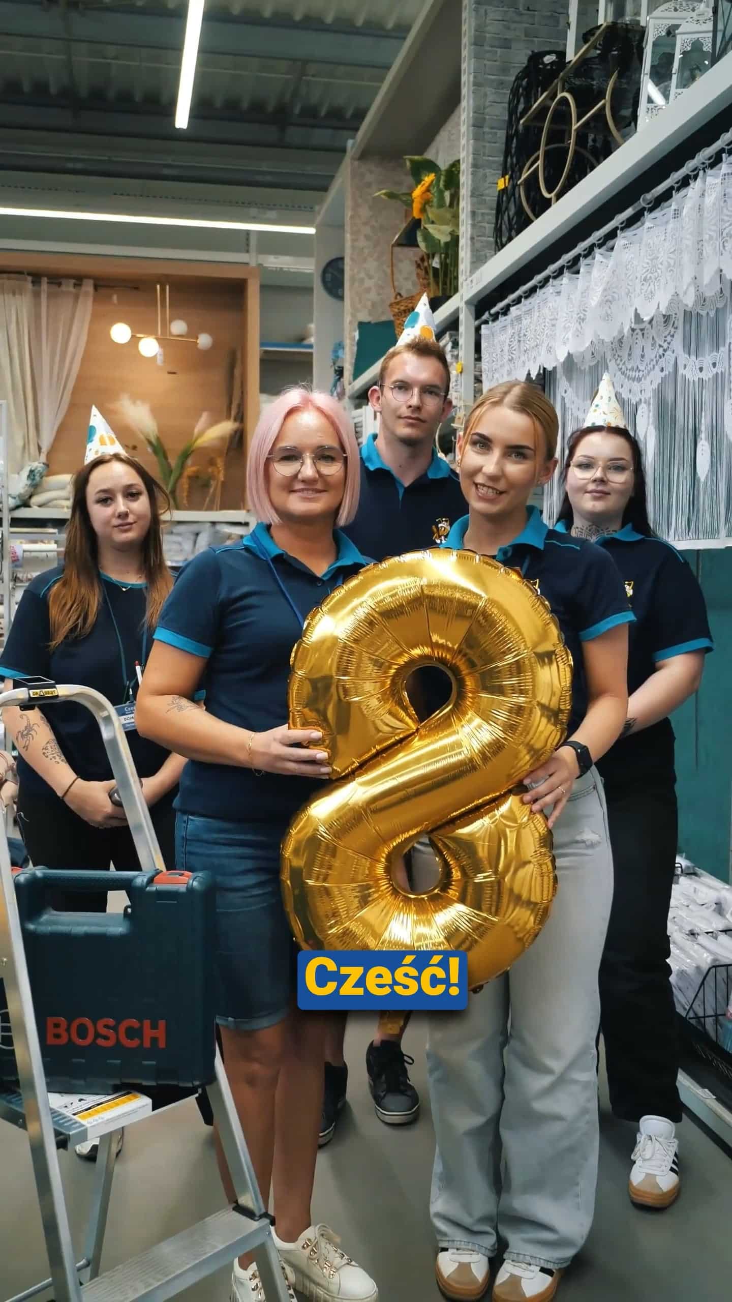 Sześć osób w imprezowych czapkach stoi razem w sklepie, dwie trzymają duży złoty balon w kształcie cyfry 8. Z przodu widnieje niebieski napis "Cześć!". (Cześć!).