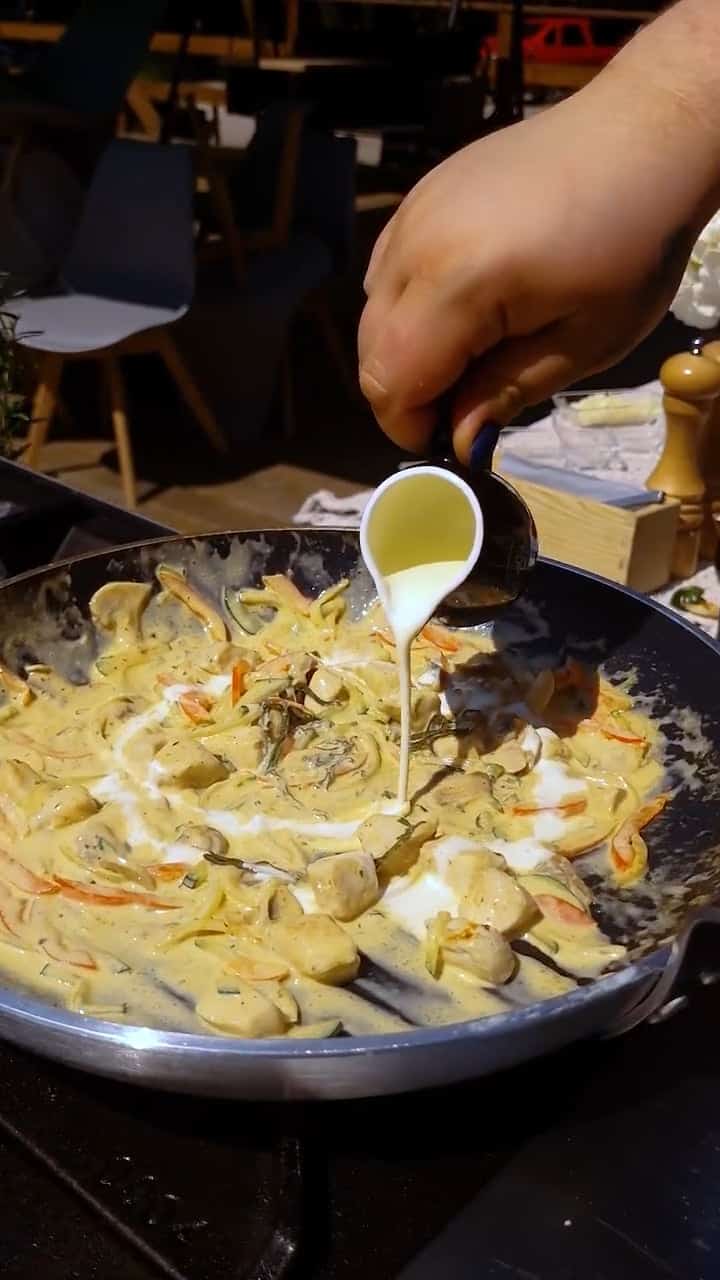 Ręka wlewa śmietanę do patelni wypełnionej kremową mieszanką grzybów i warzyw, w tle widoczne krzesła i młynek do pieprzu.