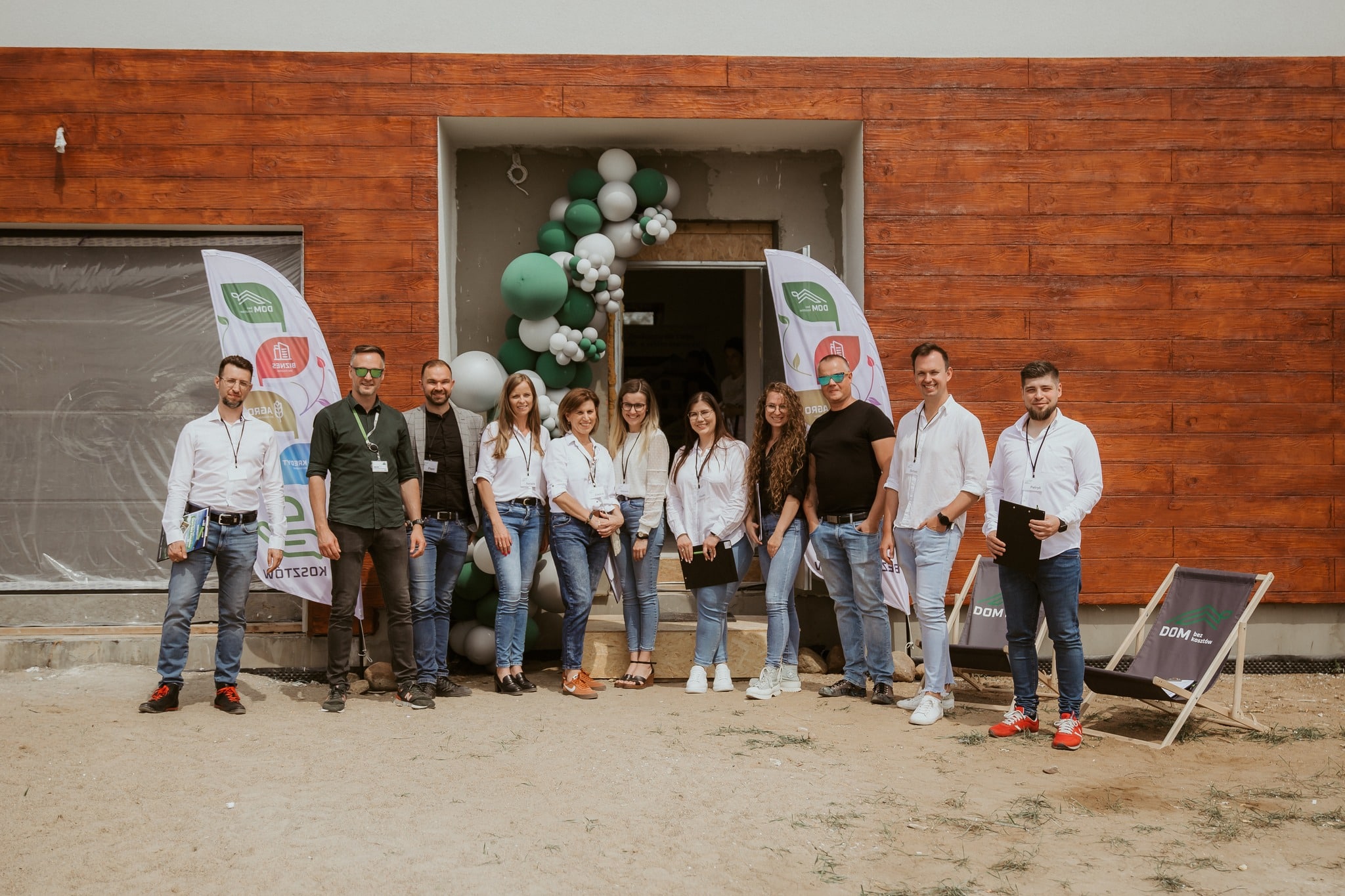 Grupa jedenastu osób stoi przed wejściem do budynku udekorowanym balonami i banerami, pozując do zdjęcia grupowego.