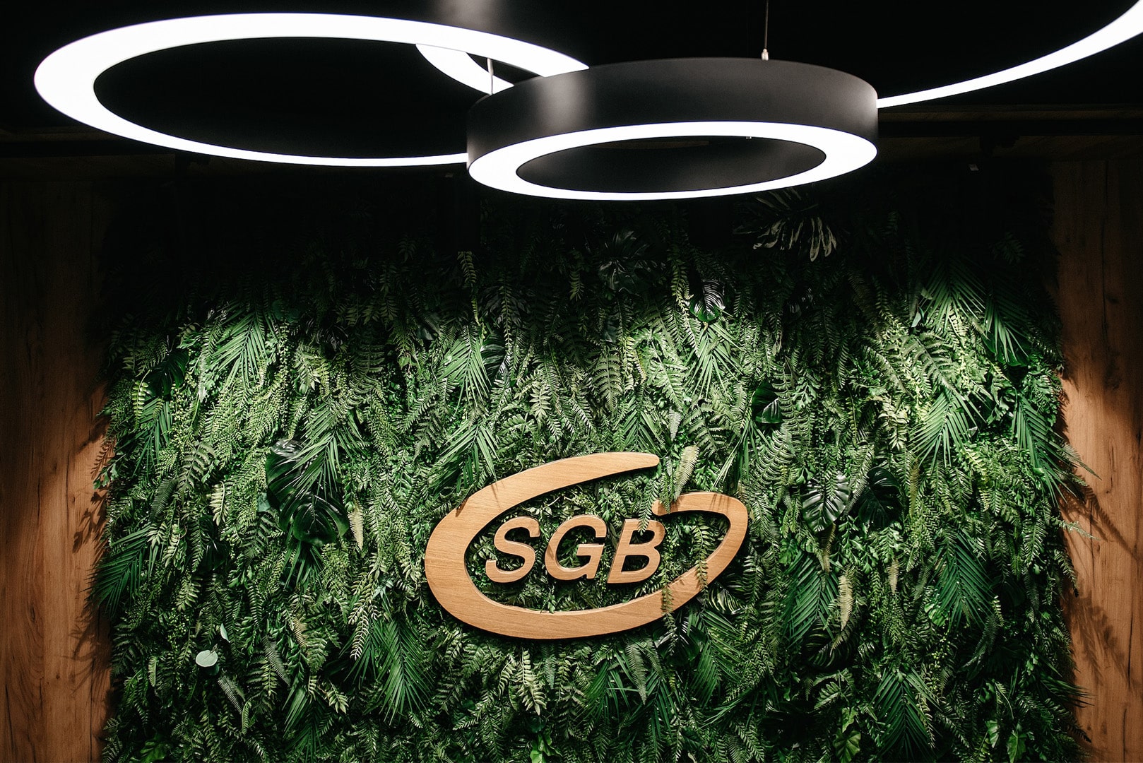 Drewniane logo "SGB" jest zamontowane na zielonej ścianie sztucznych roślin pod okrągłymi lampami sufitowymi.